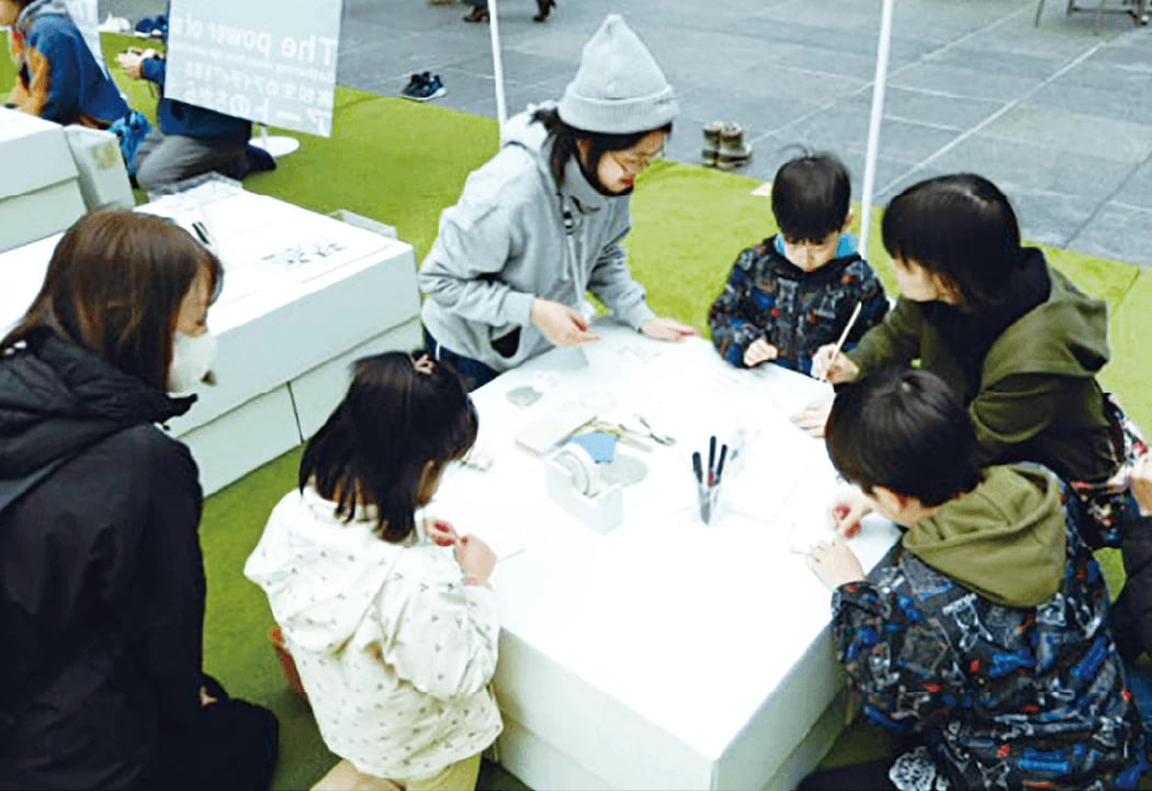 ▲エンボス加工体験の様子。高校生のスタッフはお揃いのパーカーとニット帽を身に着け、来場者をサポートする。